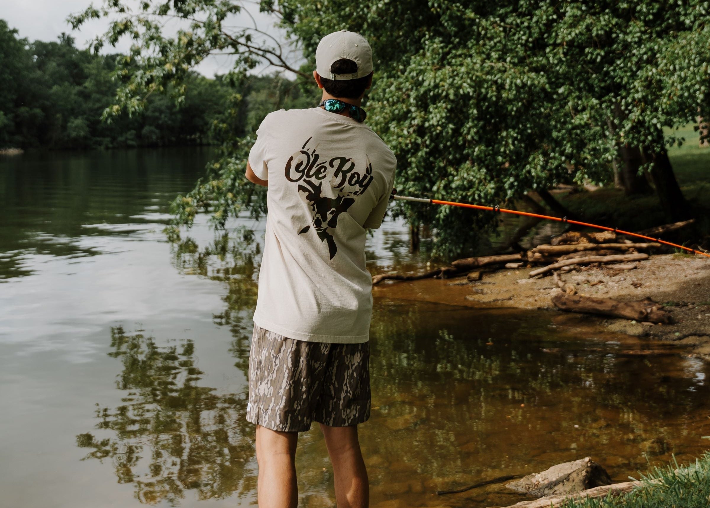 Person fishing by a lake wearing Ole Boy apparel with trees in the background