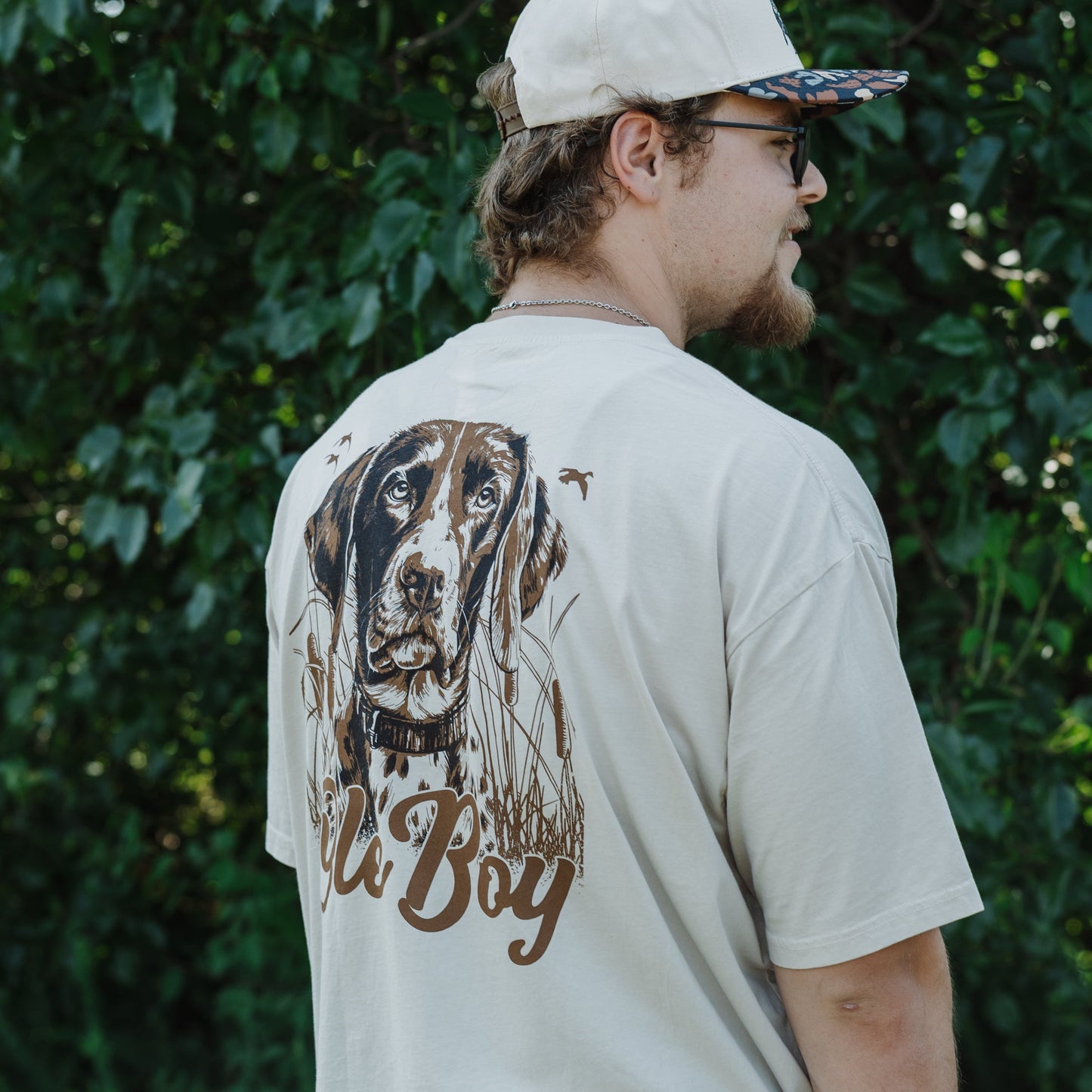 Man wearing a beige t-shirt with a GSP dog graphic and text, standing outdoors against green foliage.