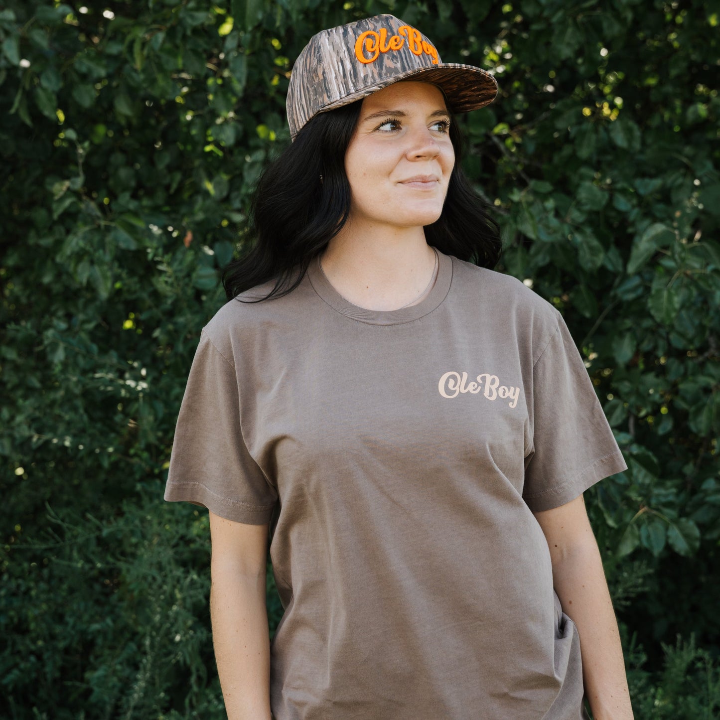 Woman wearing a brown t-shirt with 'Ole Boy' logo and a camouflage cap outdoors.