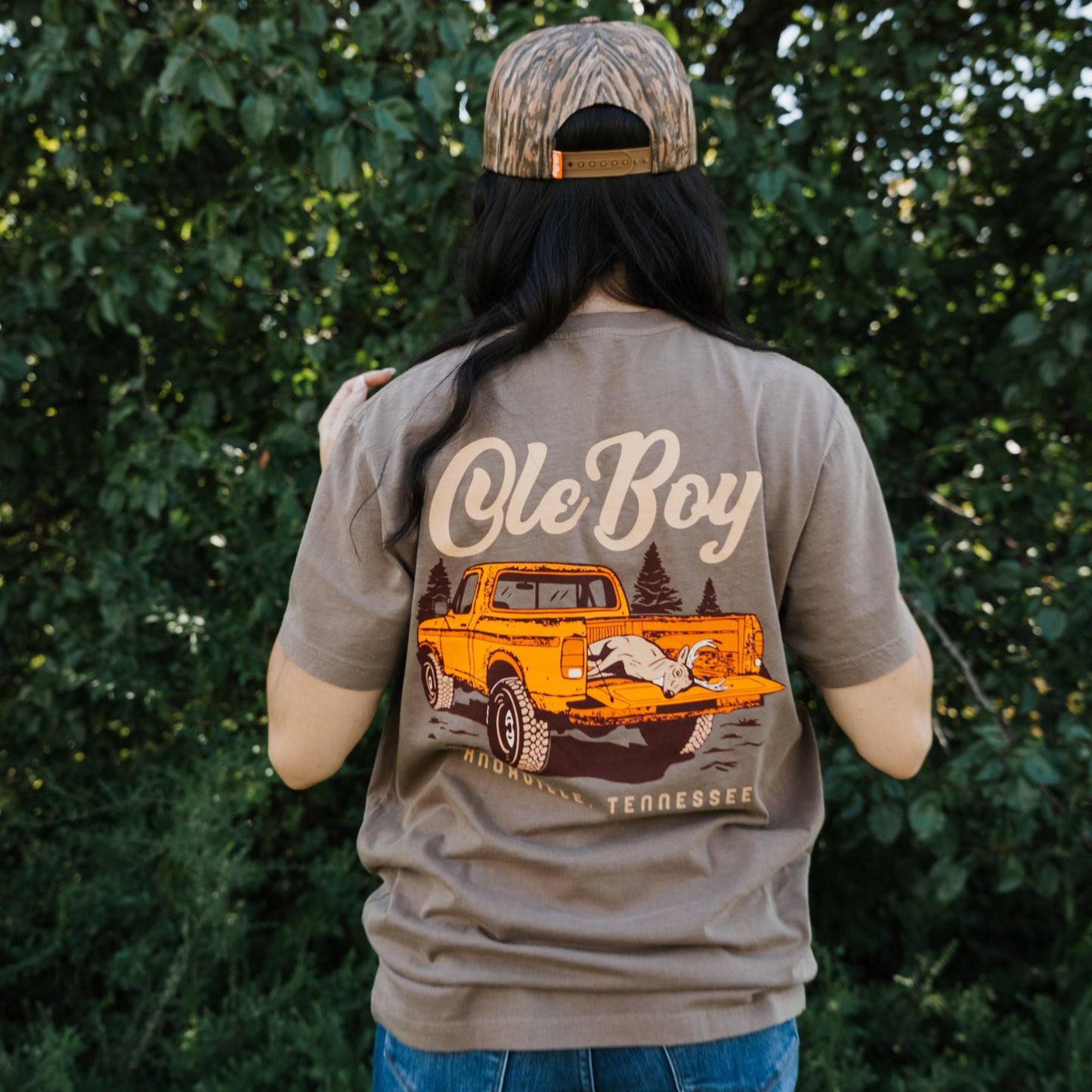 Person wearing a t-shirt with a graphic design and text, standing in front of trees.