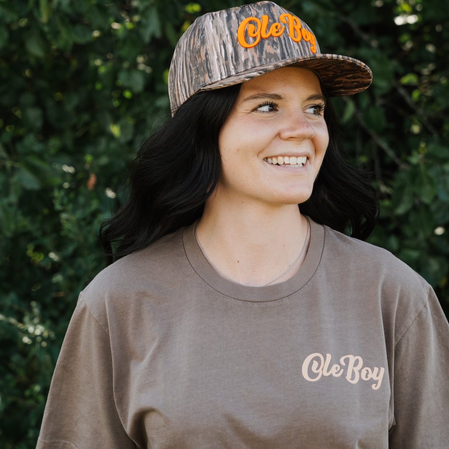 Woman wearing a camouflage cap and brown t-shirt with 'Ole Boy' branding against a green leafy background
