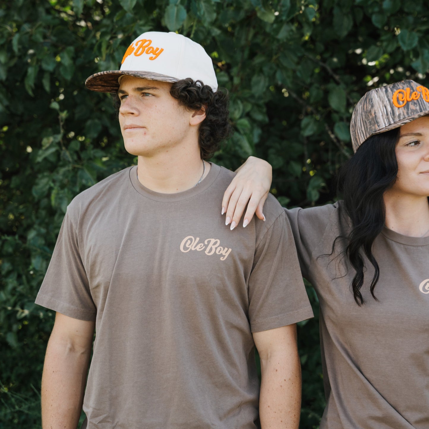 Two people wearing brown 'Ole Boy' t-shirts and caps outdoors.