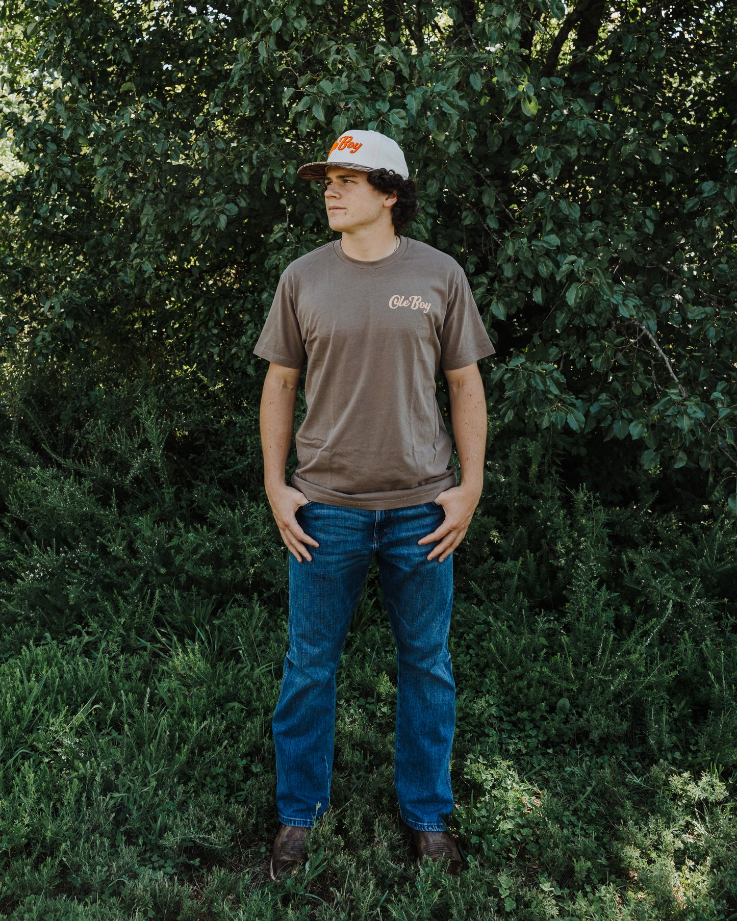 Man wearing a brown t-shirt and blue jeans standing in a forest