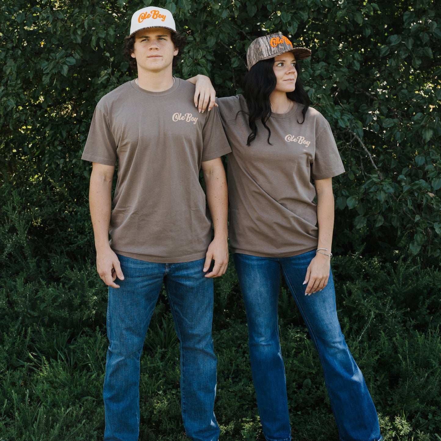Two people wearing matching brown t-shirts and jeans standing in a forest.