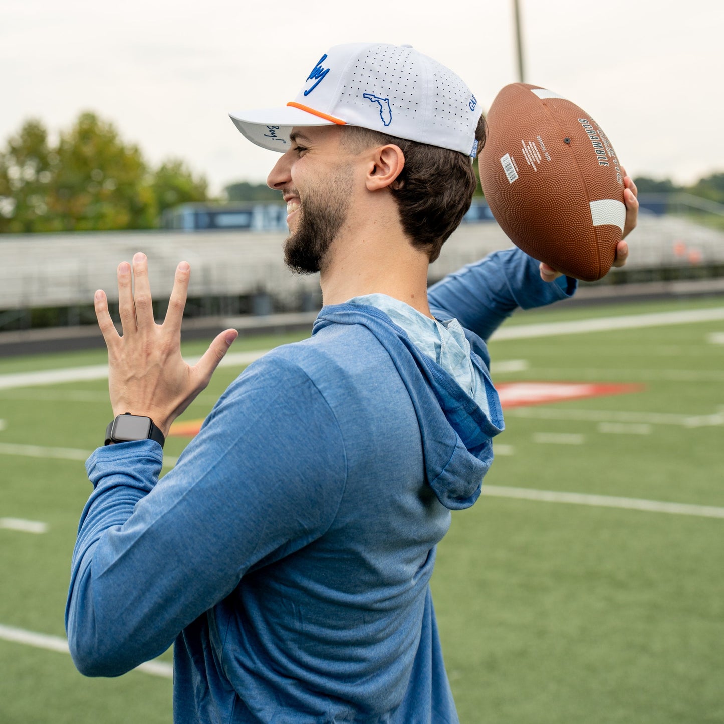 Man on a football field wearing a blue hoodie and cap, holding a football.