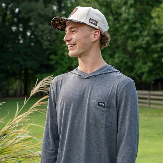 Person wearing a gray hoodie with a logo, standing outdoors with greenery in the background