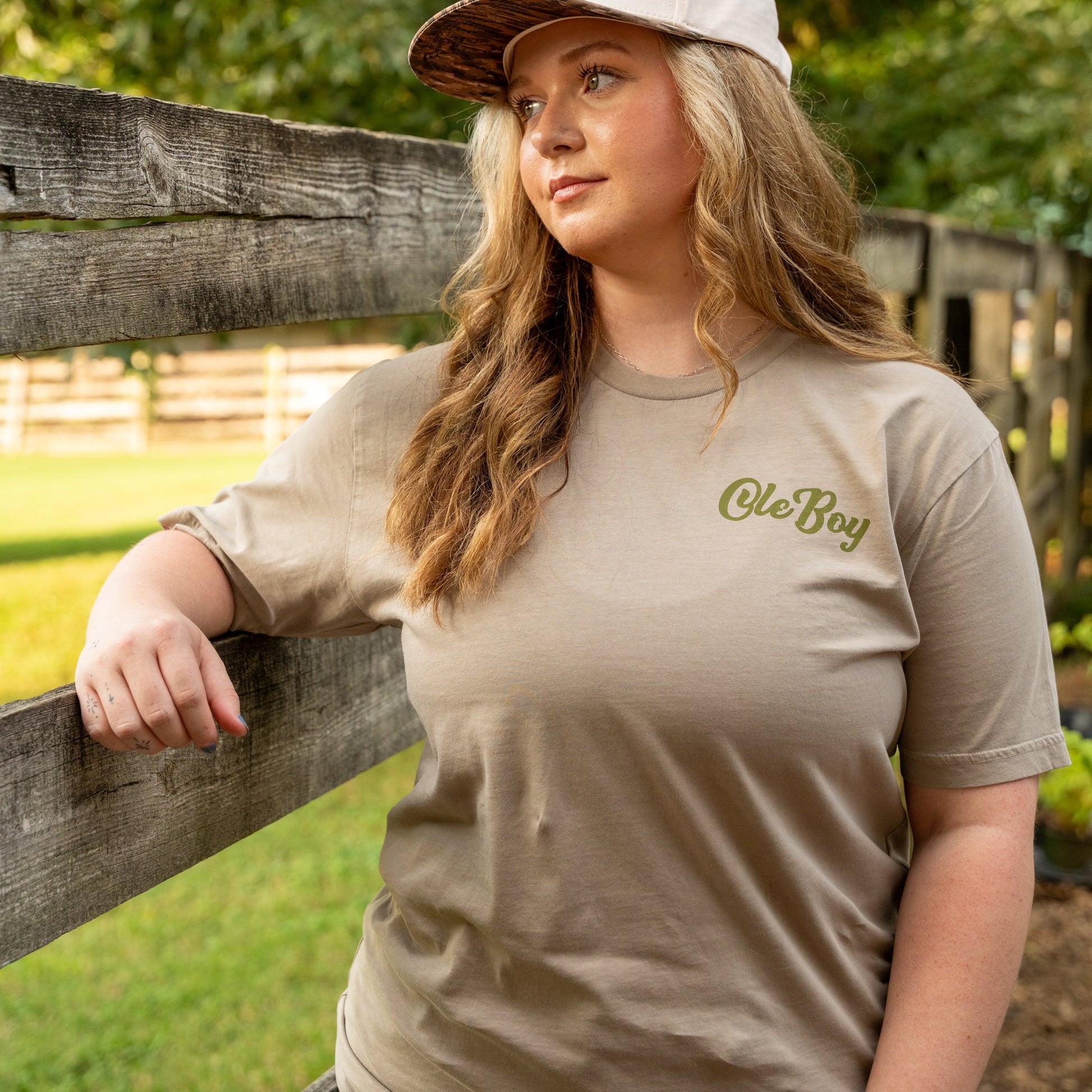 Woman wearing a beige t-shirt and cap with 'Ole Boy' branding, standing outdoors.