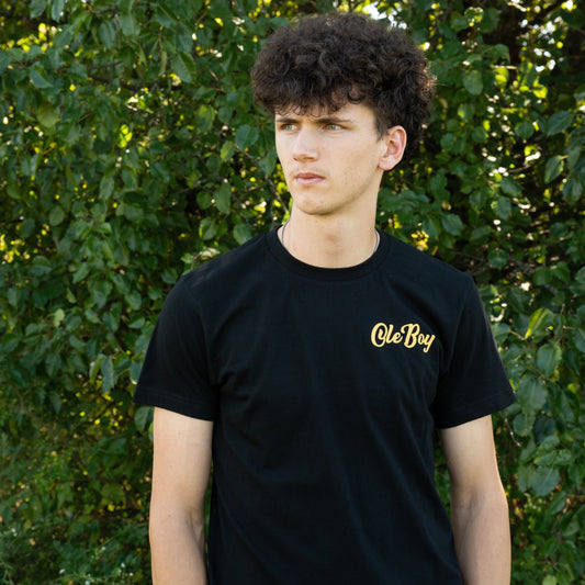 Person wearing a black t-shirt with 'Ole Boy' logo in front of green foliage