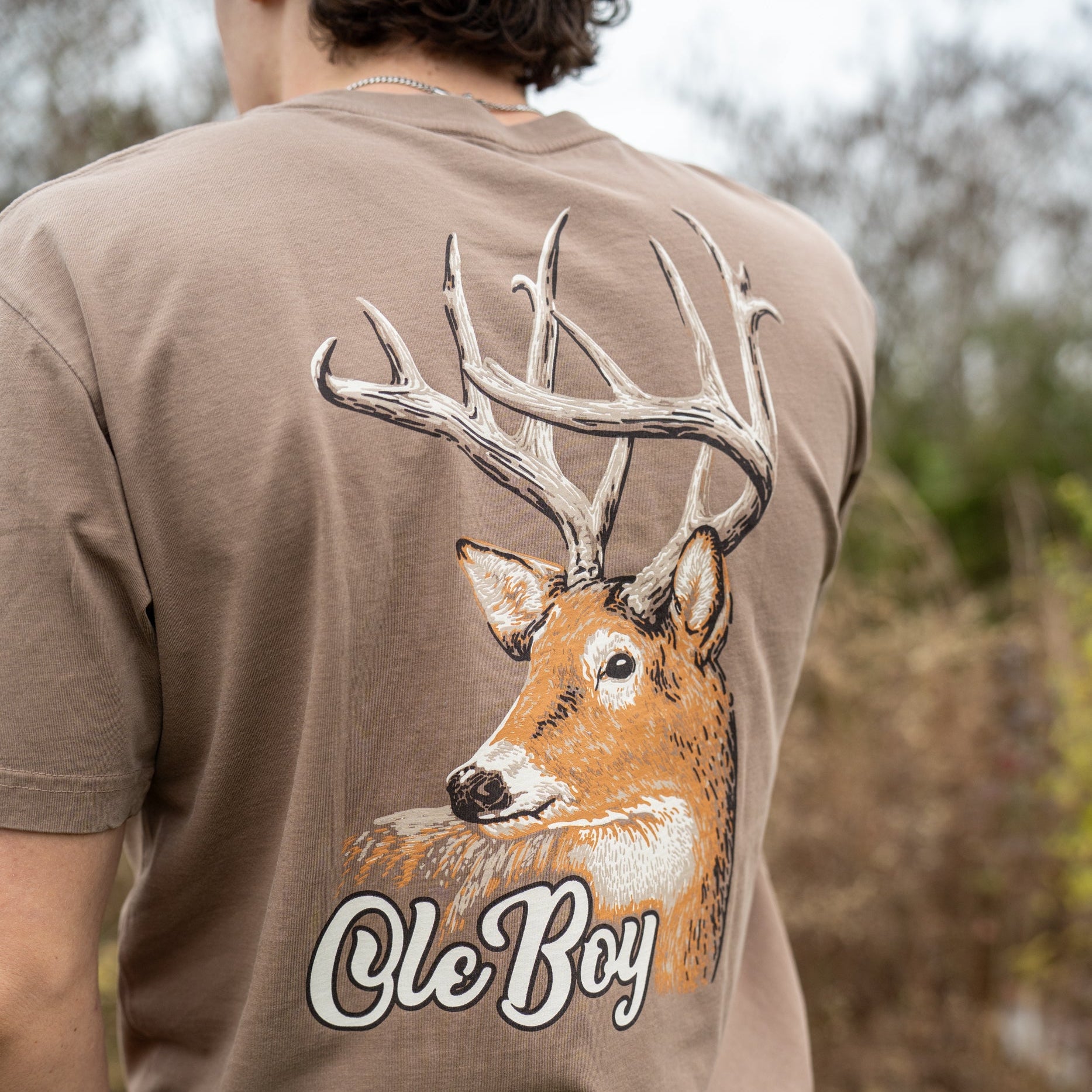Person wearing a brown t-shirt with a deer graphic and 'Ole Boy' text in an outdoor setting