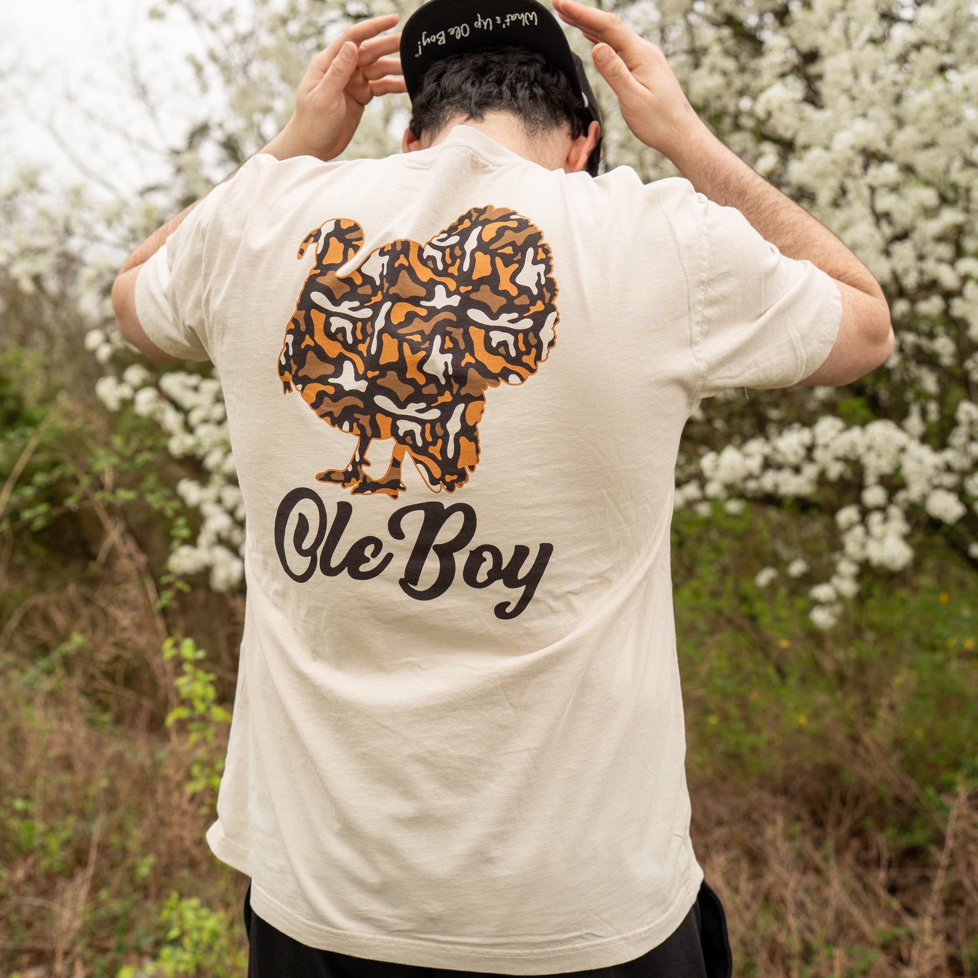 Person wearing a white t-shirt with a graphic and text, standing in a natural setting with trees in the background.