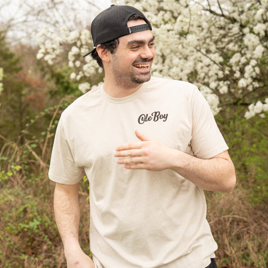 Man wearing a beige t-shirt with 'Ole Boy' logo outdoors among trees with white blossoms.