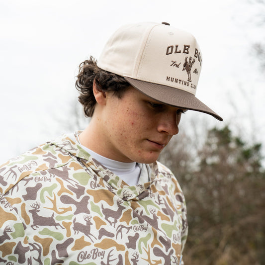 Person wearing a camouflage jacket and a cap with 'OLE BOY' branding outdoors.