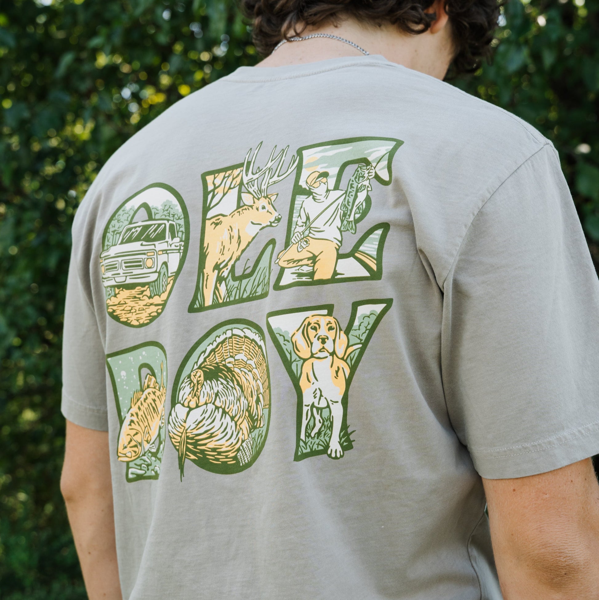 Person wearing a t-shirt with a graphic design on the back, standing in front of green foliage.