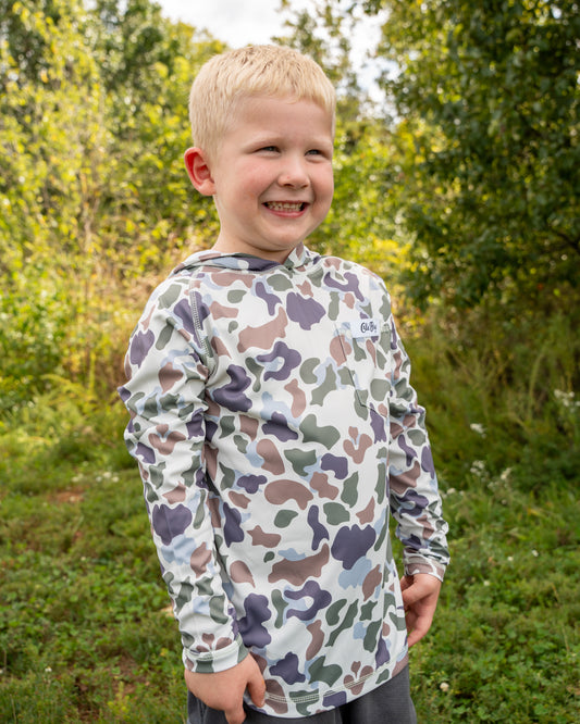 Child wearing a camouflage-patterned shirt outdoors with greenery in the background