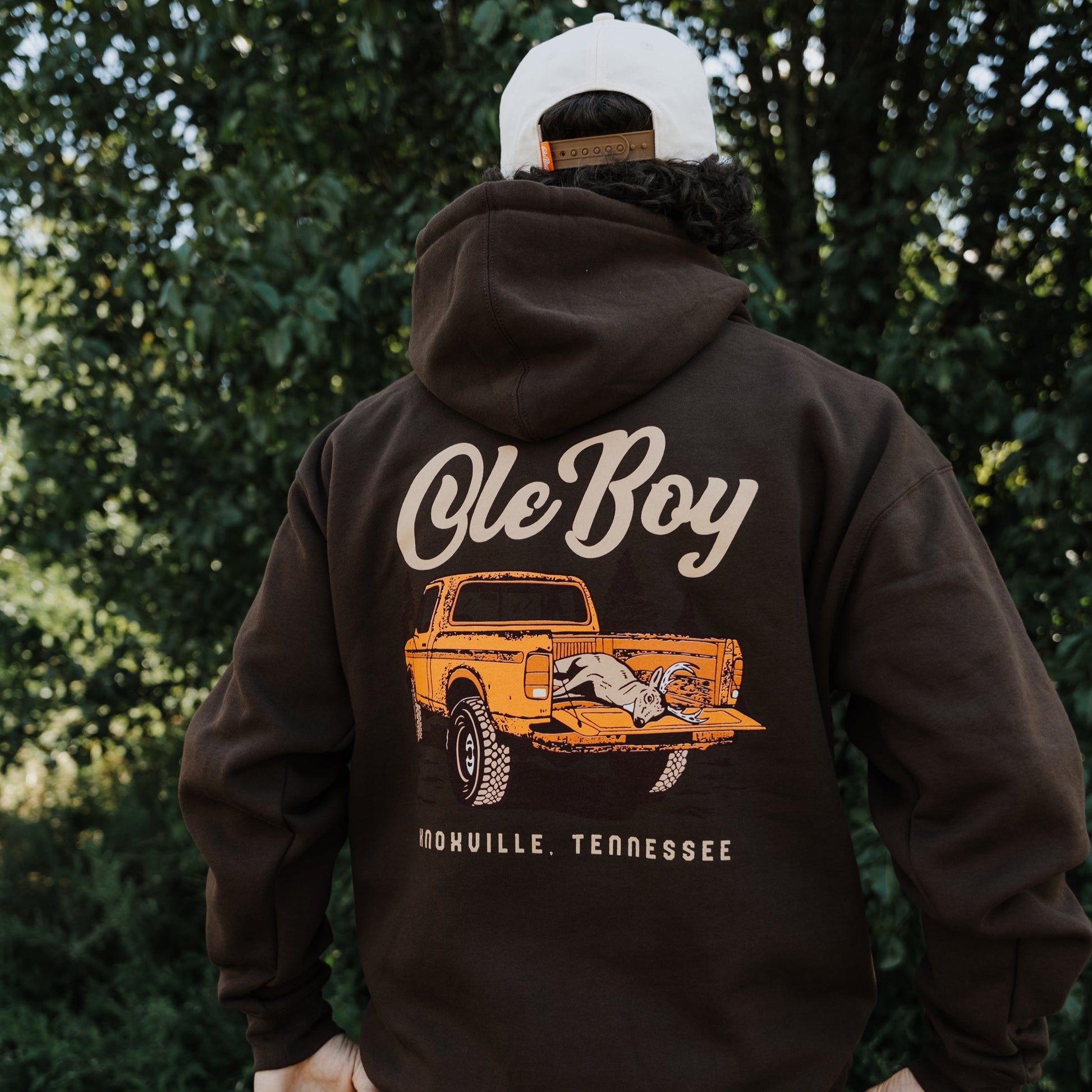 Person wearing a brown hoodie with 'Ole Boy' and truck graphic, standing outdoors.