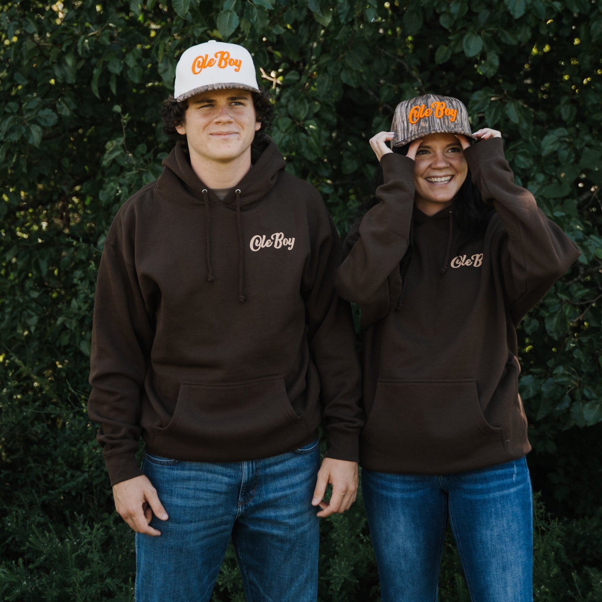 Two people wearing brown hoodies and hats with a logo, standing in front of green foliage.