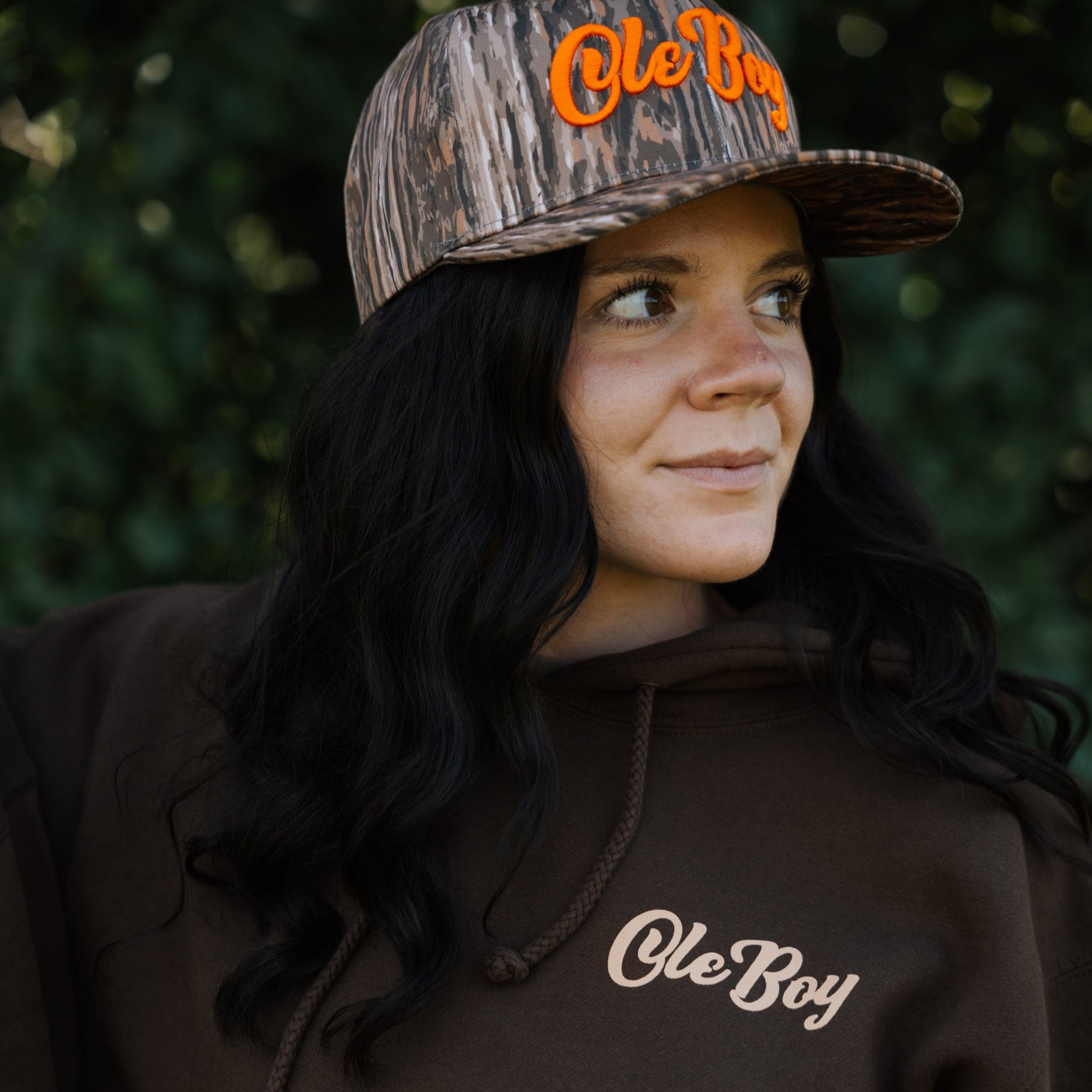 Person wearing a brown hoodie and cap with 'Ole Boy' branding against a blurred green background