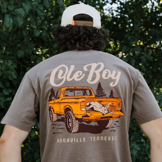 Person wearing a brown t-shirt with a graphic of a truck and 'Ole Boy' text, standing against a green leafy background.