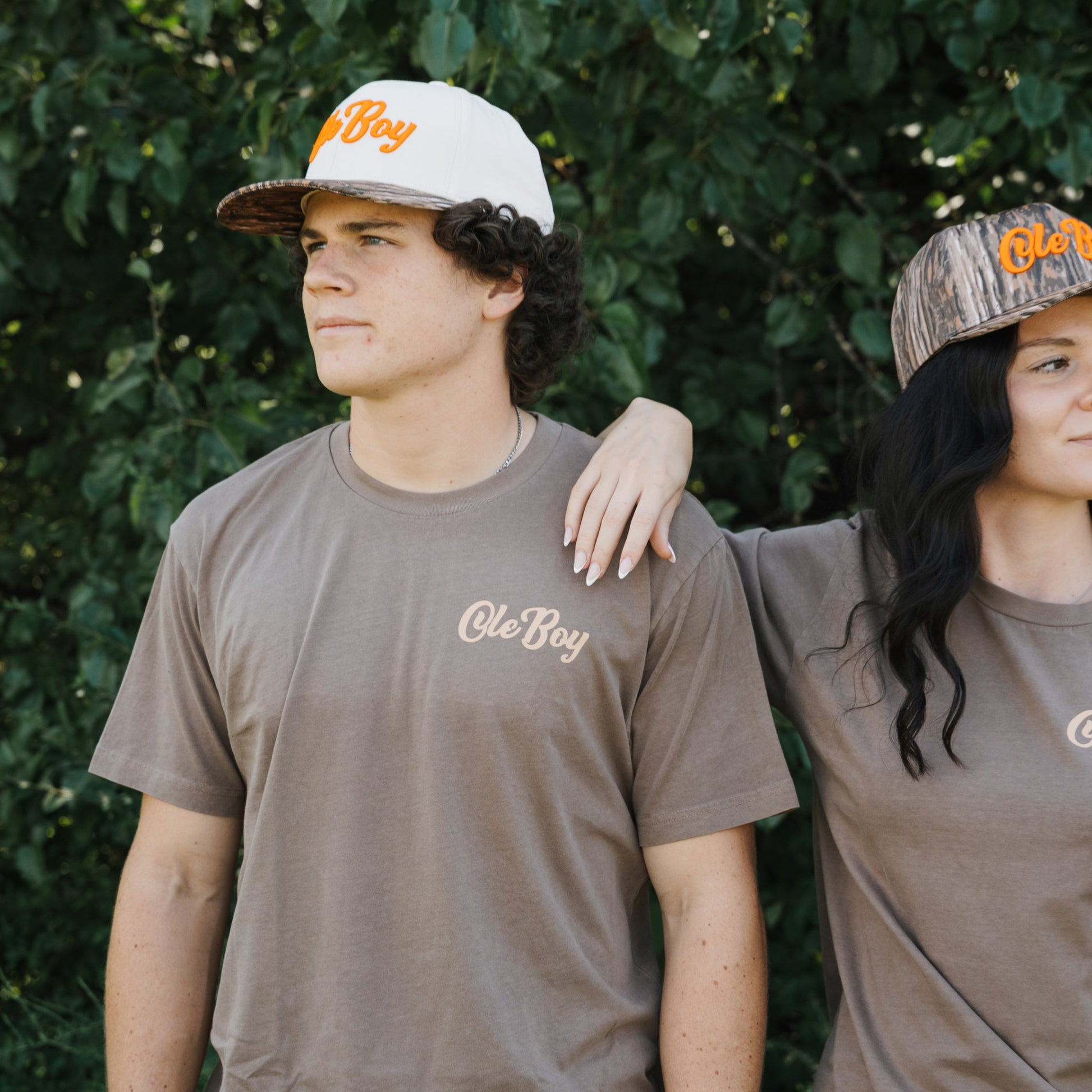 Two people wearing brown 'Ole Boy' t-shirts and caps outdoors.