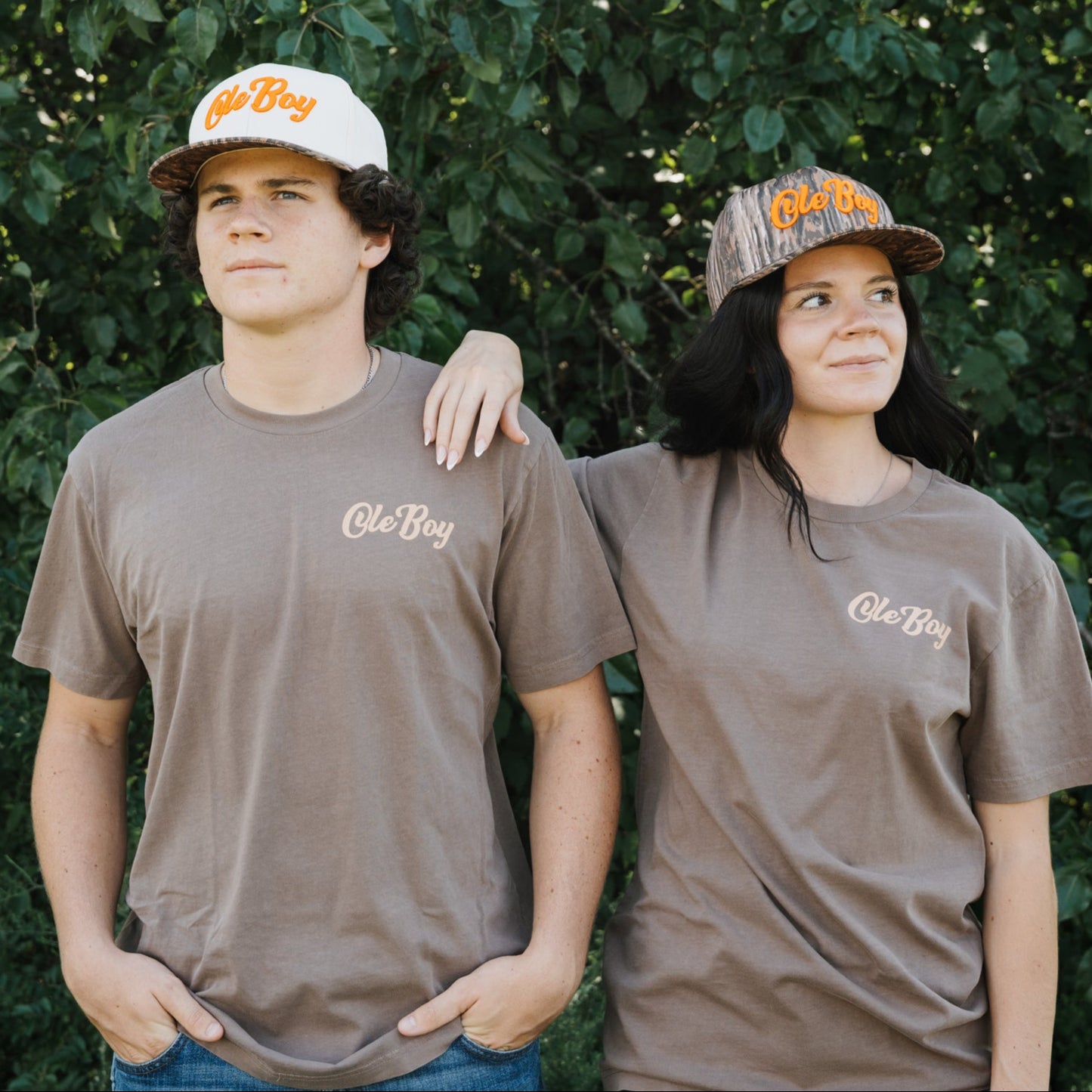 Two people wearing matching brown t-shirts and hats in front of a green leafy background