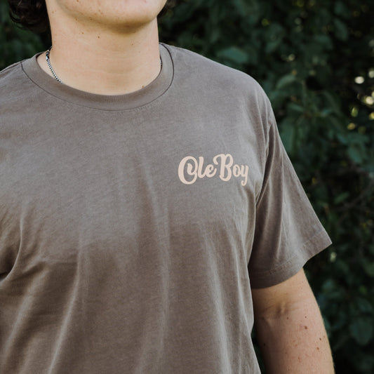 Person wearing a brown t-shirt with 'Ole Boy' text against a blurred green background