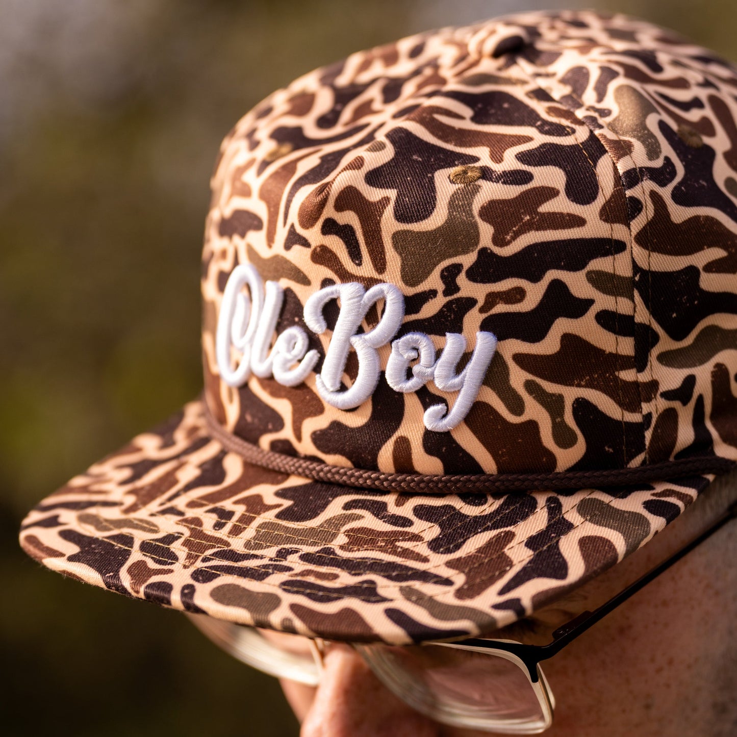 Camouflage-patterned cap with 'Ole Boy' text held by a person against a blurred natural background