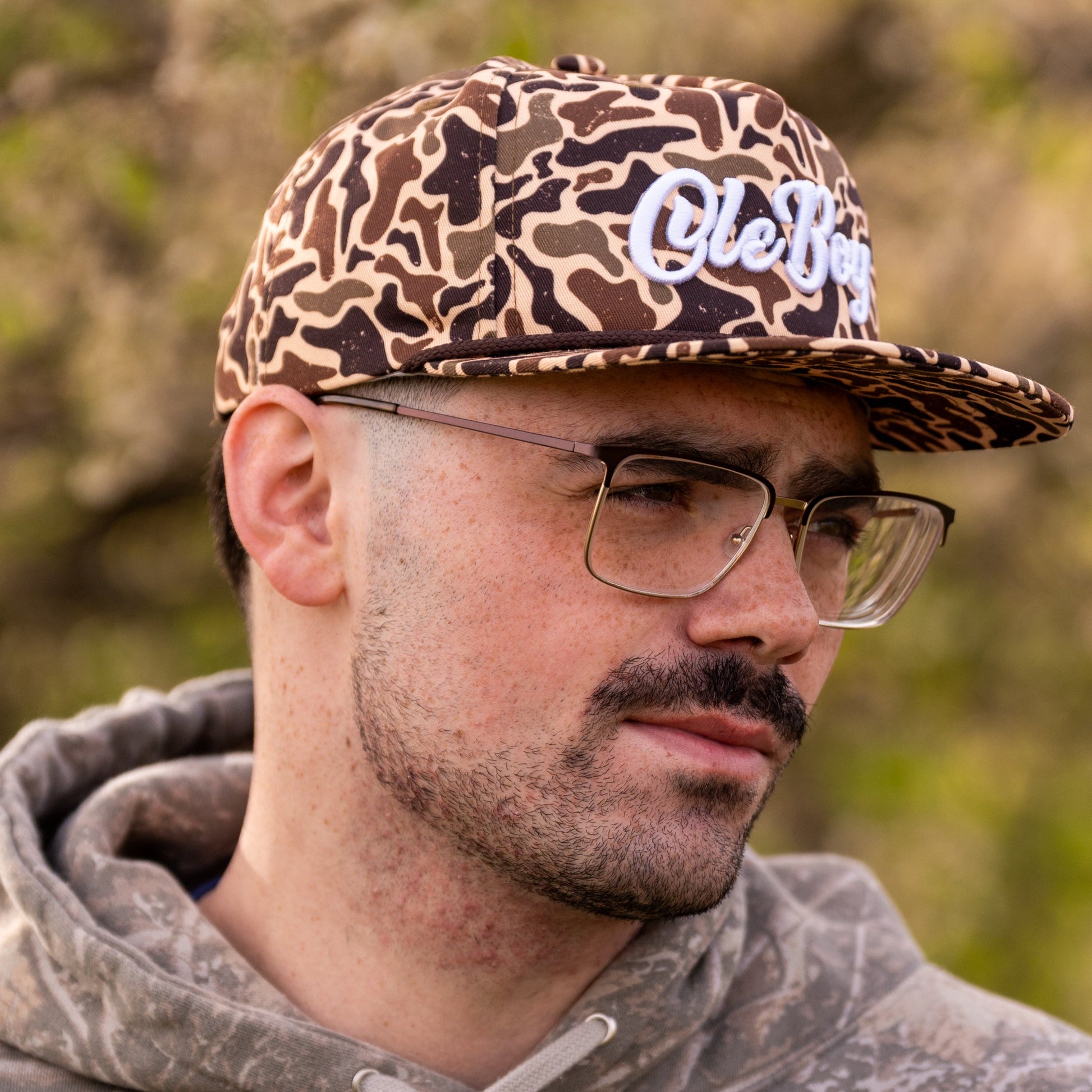 Man wearing a camouflage cap with 'Ole Boy' branding and a gray hoodie outdoors.