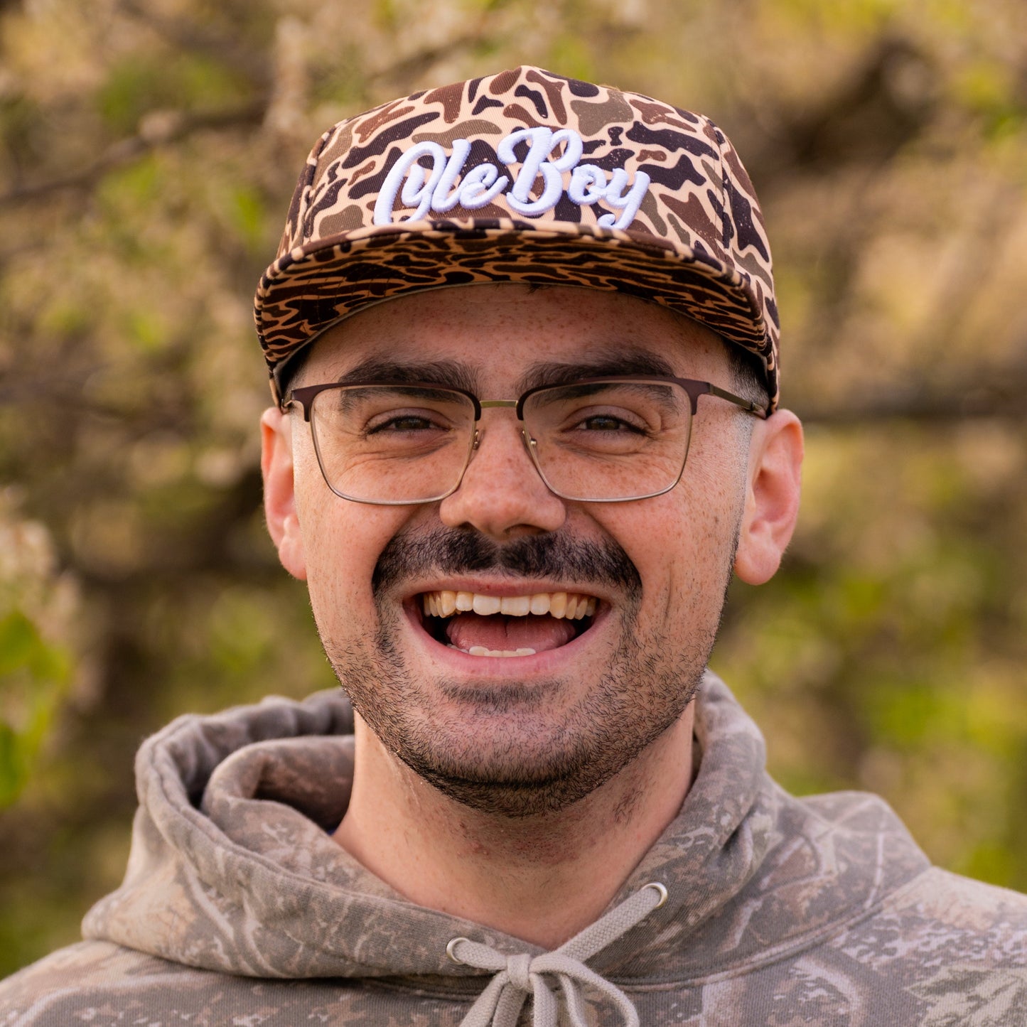 Man wearing a camouflage hoodie and leopard print cap with 'Ole Boy' text, outdoors.