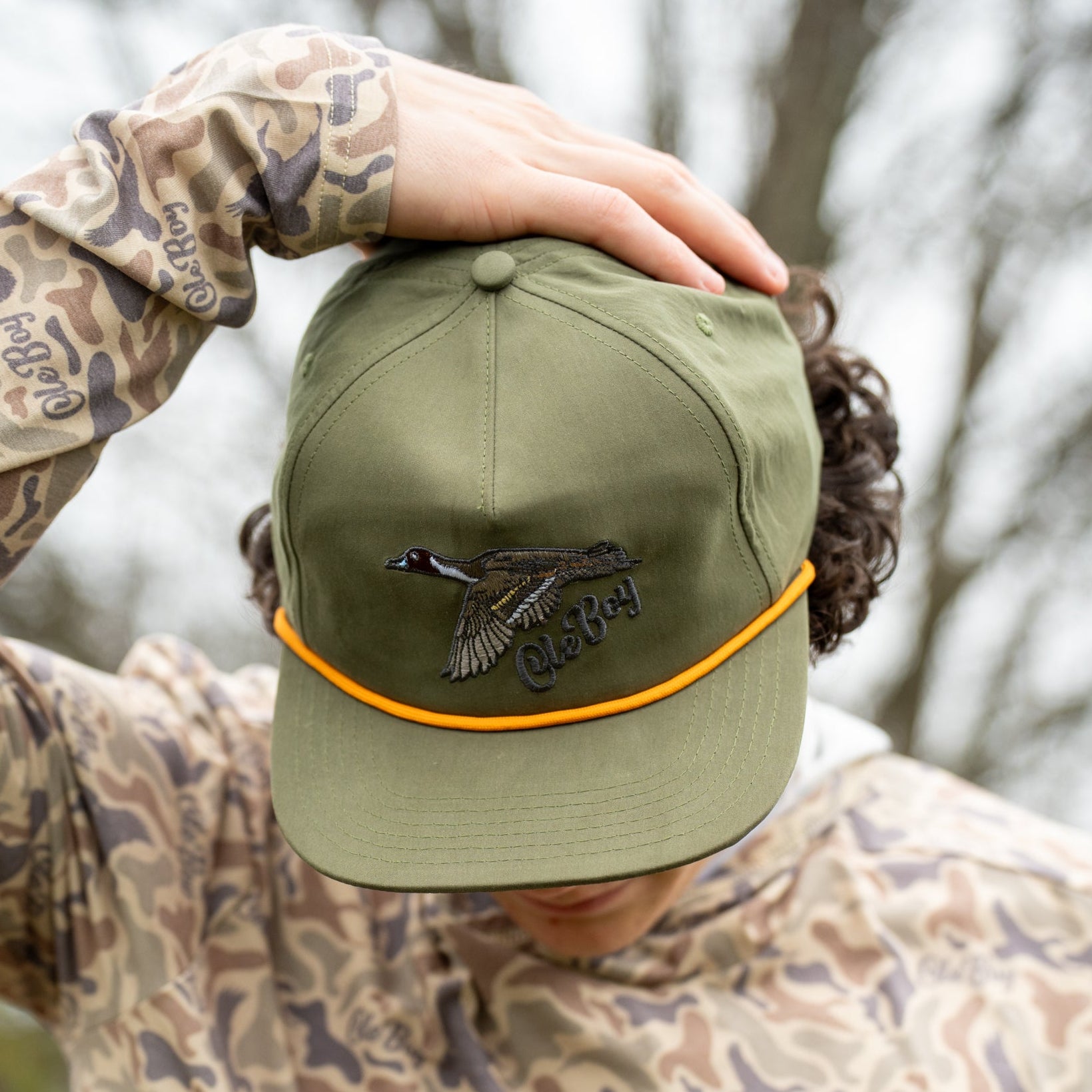 Person wearing a green cap with a logo and camouflage jacket outdoors