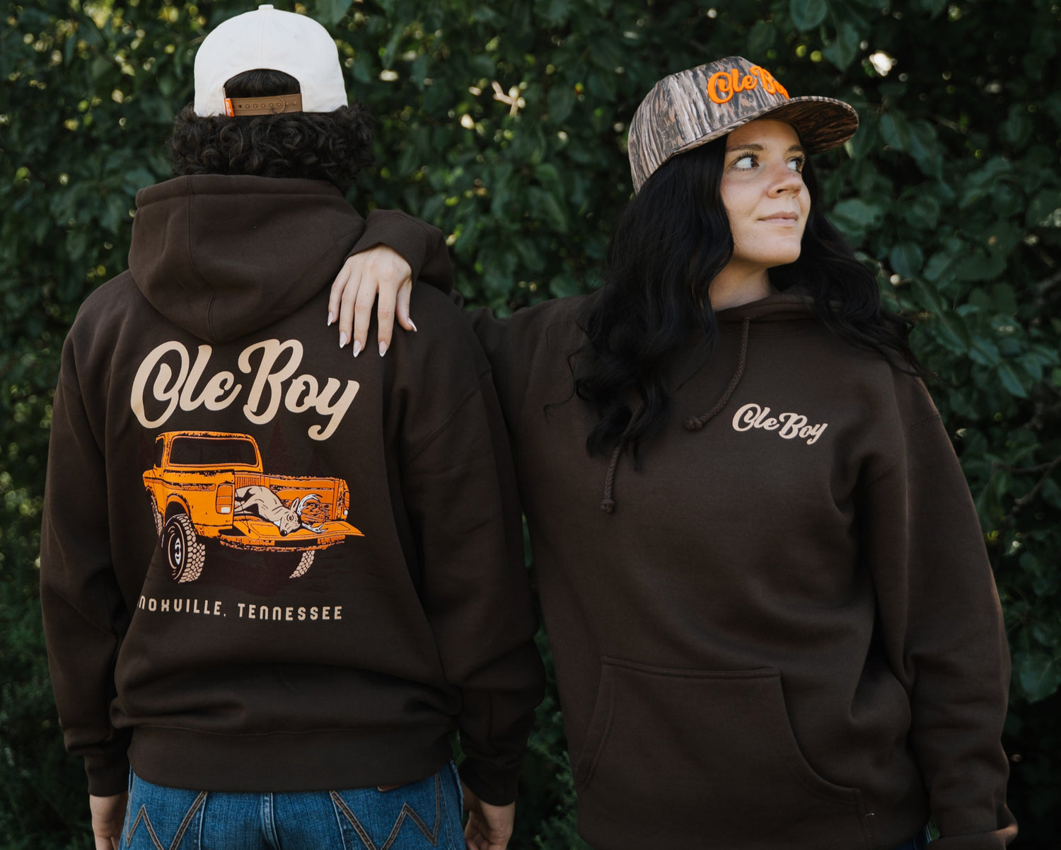 Two people wearing brown hoodies with 'Ole Boy' branding, one with a graphic of a truck on the back, against a green leafy background.