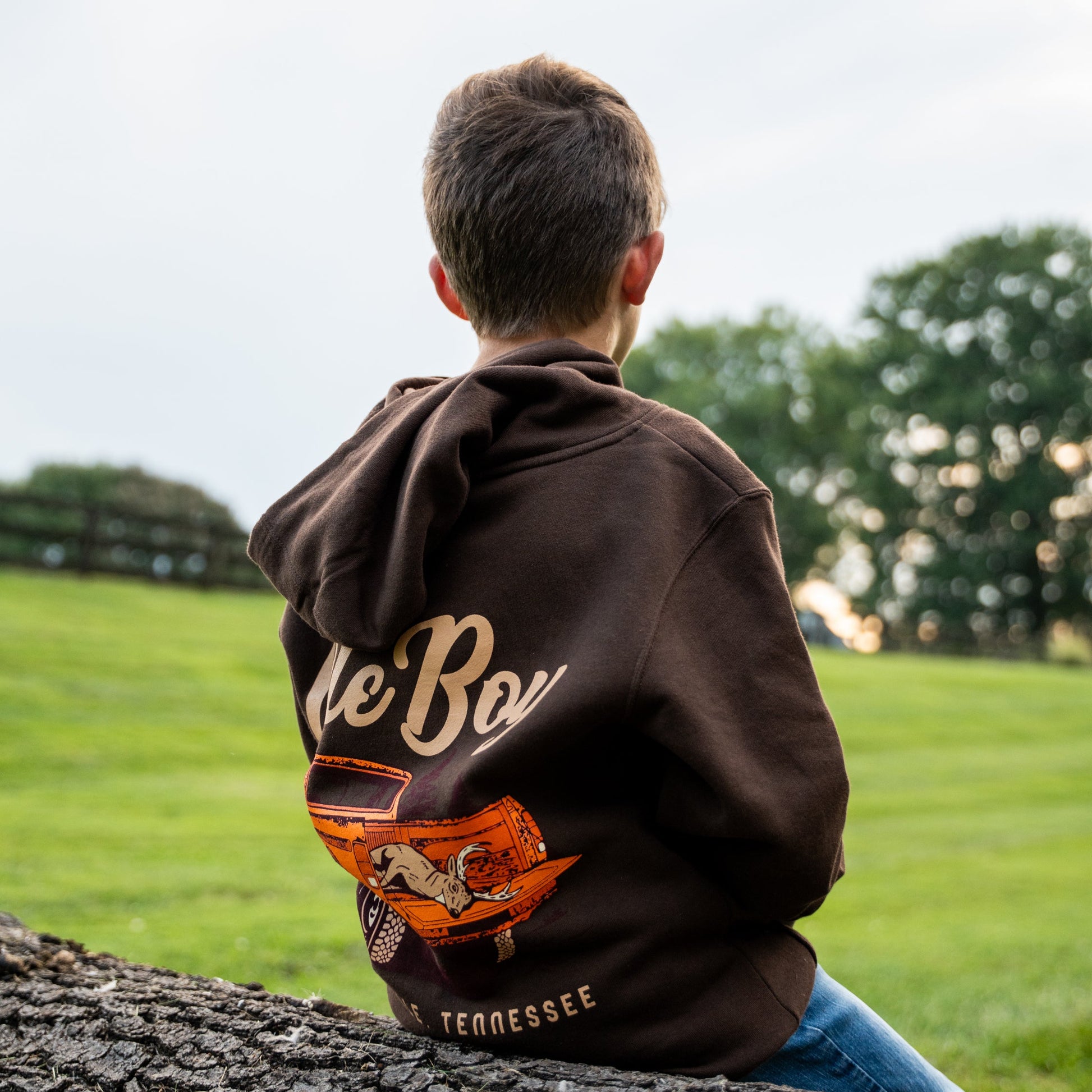Child wearing a hoodie with 'Ole Boy' branding sitting on a log in a grassy field.