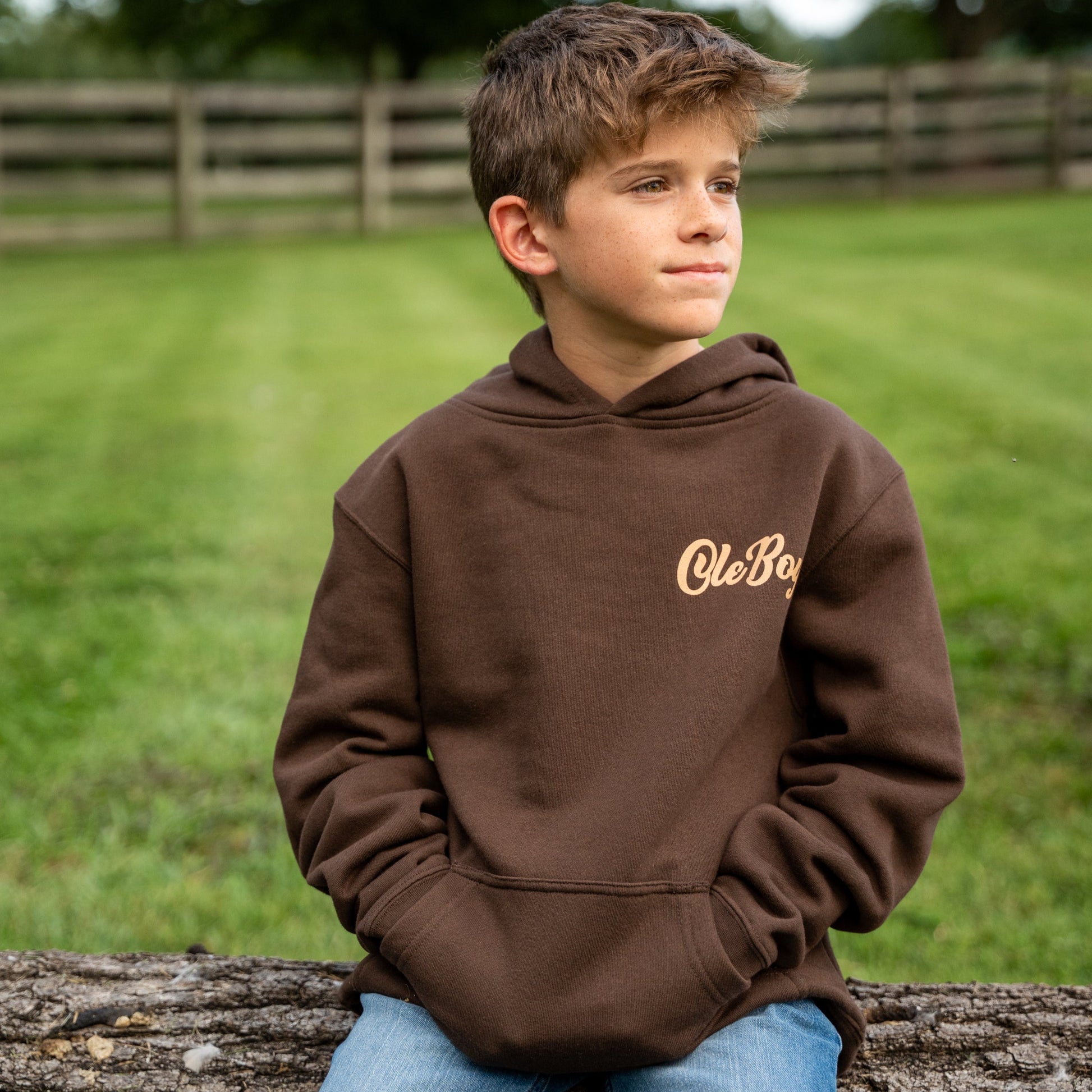 Child wearing a brown hoodie with 'Ole Boy' logo, sitting on a log outdoors.
