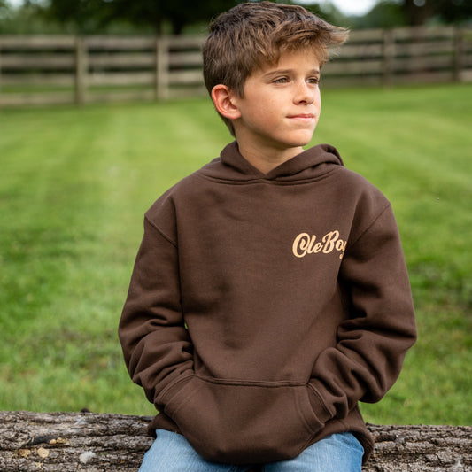 Child wearing a brown hoodie with 'Ole Boy' logo, sitting on a log outdoors.