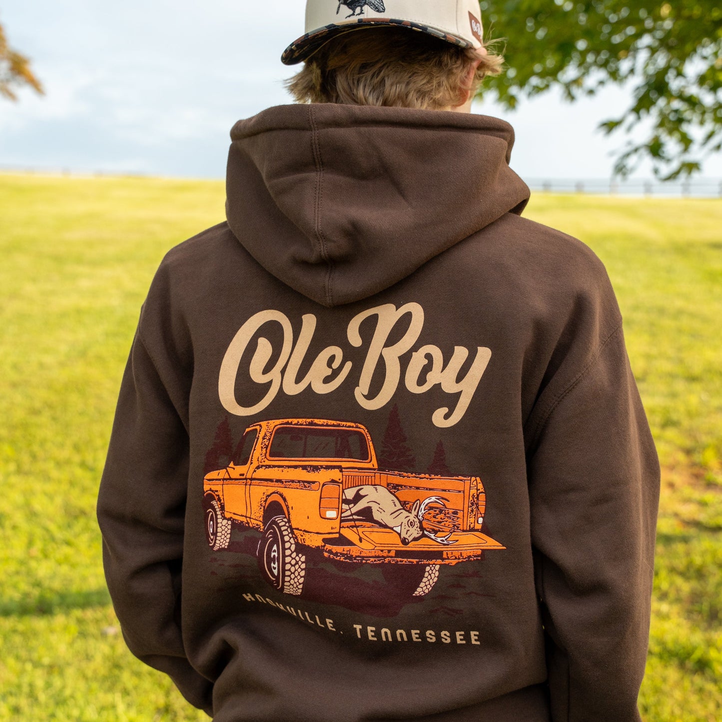 Person wearing a brown hoodie with 'Ole Boy' and truck graphic, standing in a grassy field.