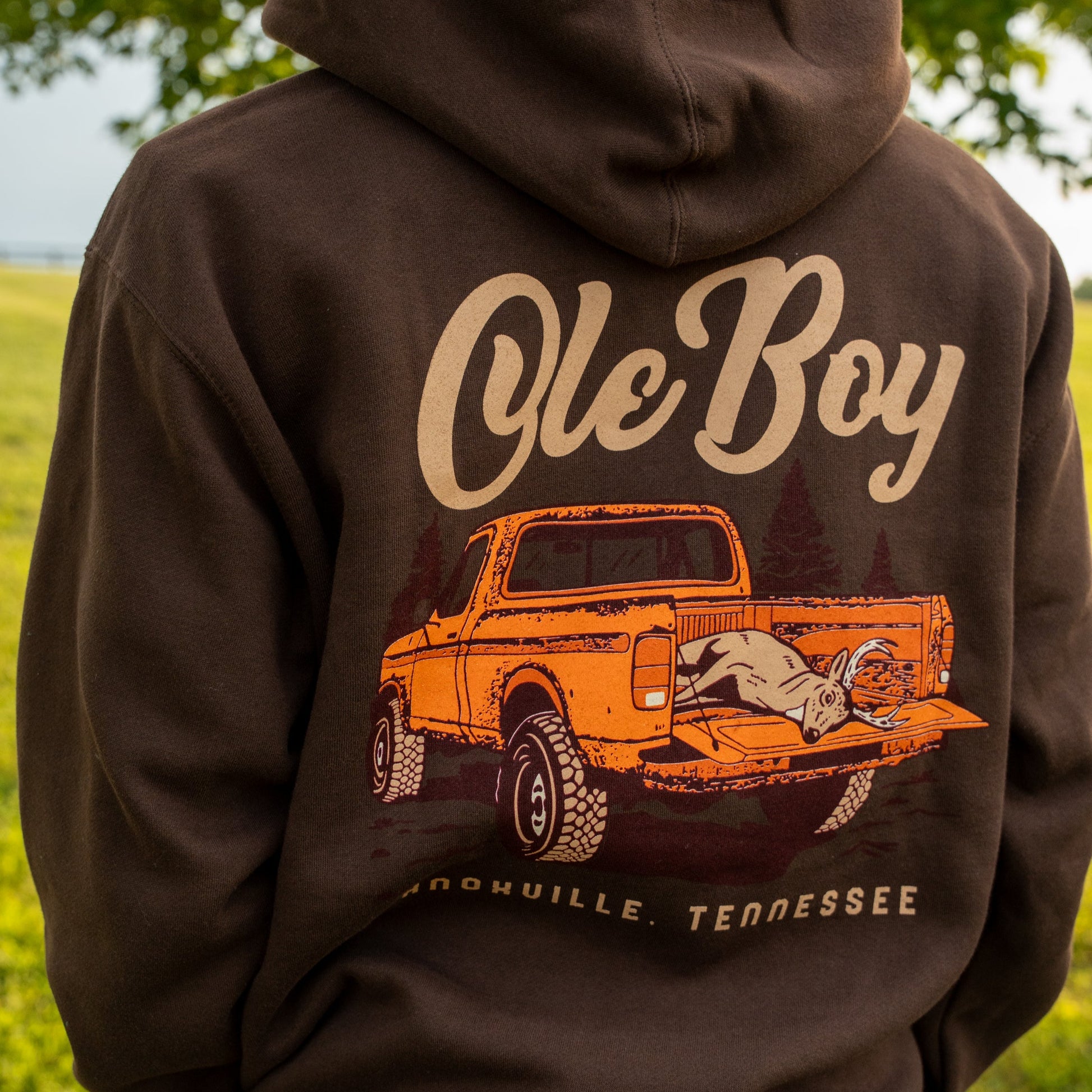 Brown hoodie with 'Ole Boy' graphic of a truck and deer on the back, outdoors.