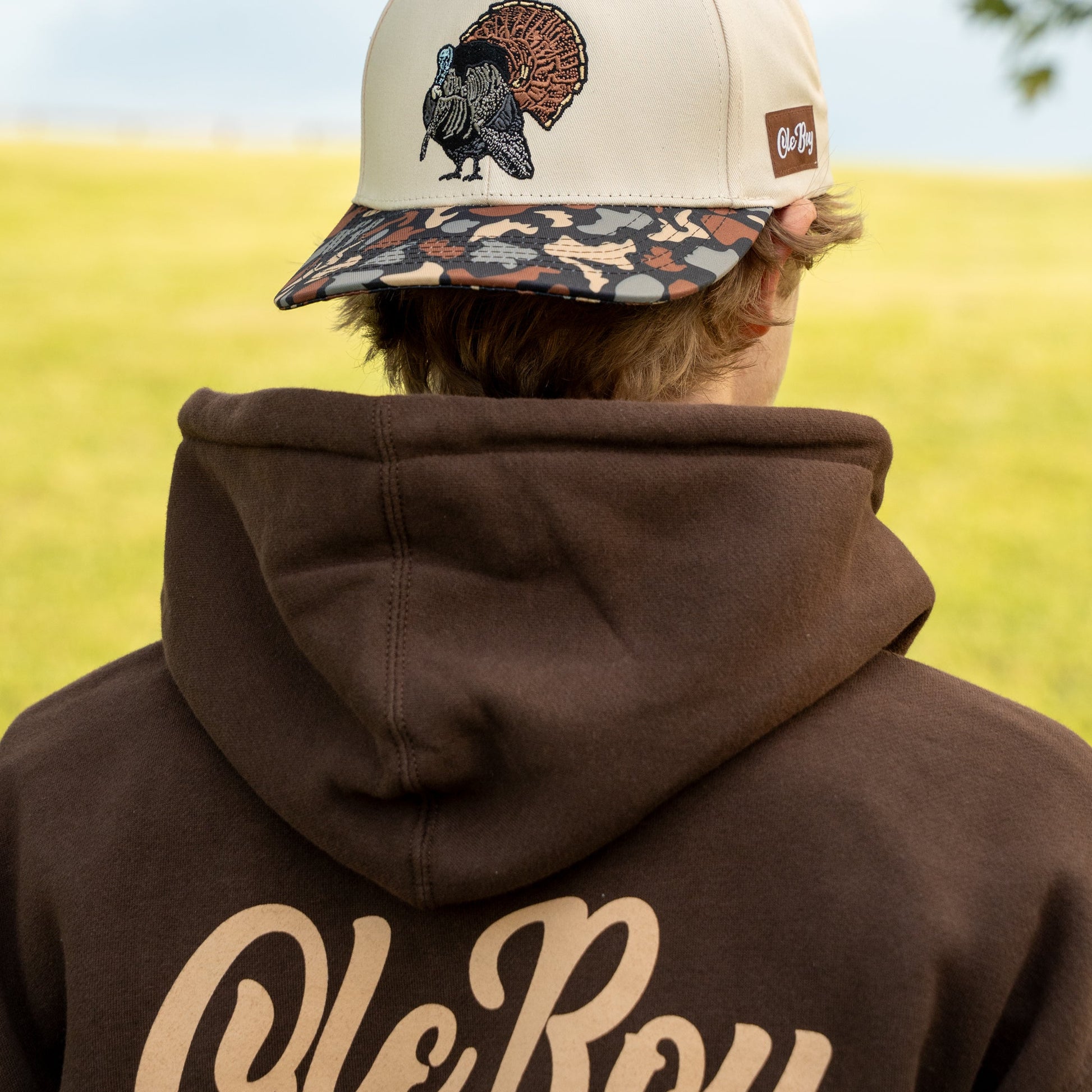 Person wearing a beige cap with a turkey design and a brown hoodie with 'Ole Boy' branding, standing in a field.