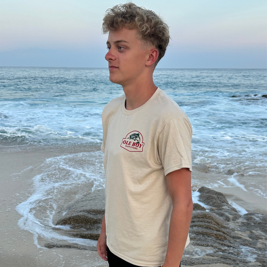 Person wearing a tan ole boy t-shirt with an ole boy logo on a beach