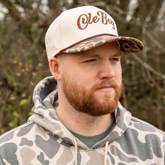 Man wearing a camouflage hoodie and cap with 'Ole Boy' logo outdoors.