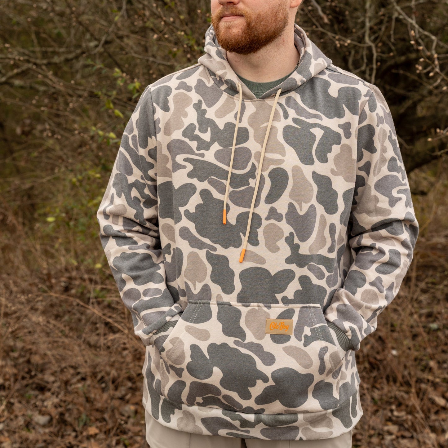 Man wearing a camouflage hoodie standing in a natural setting with trees and dry leaves.