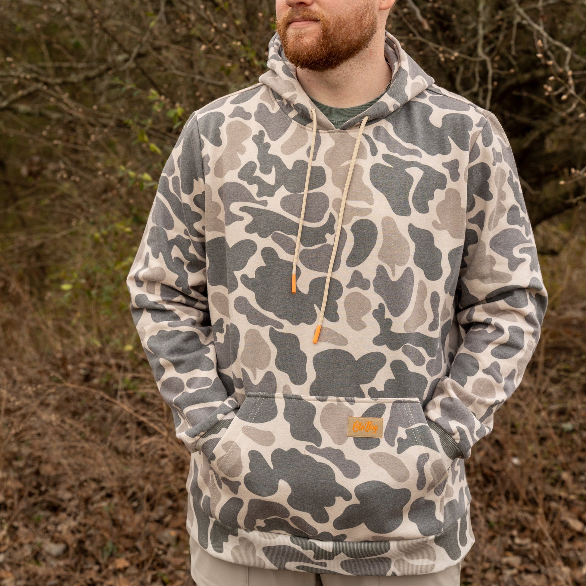 Man wearing a camouflage hoodie standing in a natural setting with trees and dry leaves.