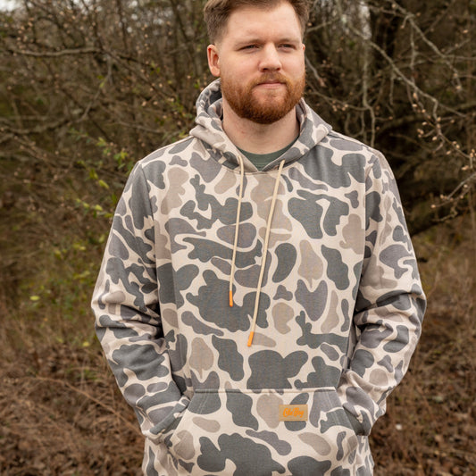 Man wearing a camouflage hoodie standing outdoors with trees in the background
