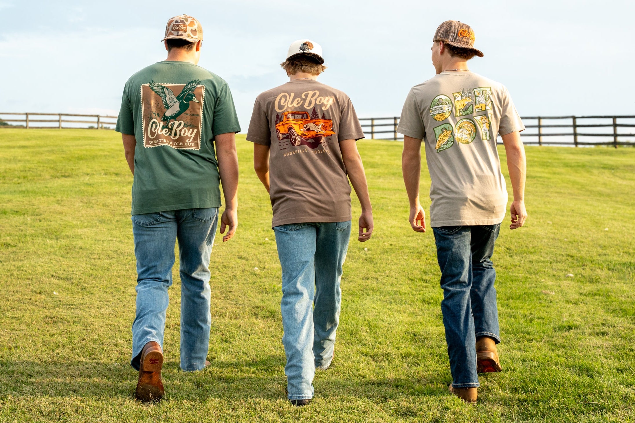 Three men walking on a grassy field wearing t-shirts with branding.