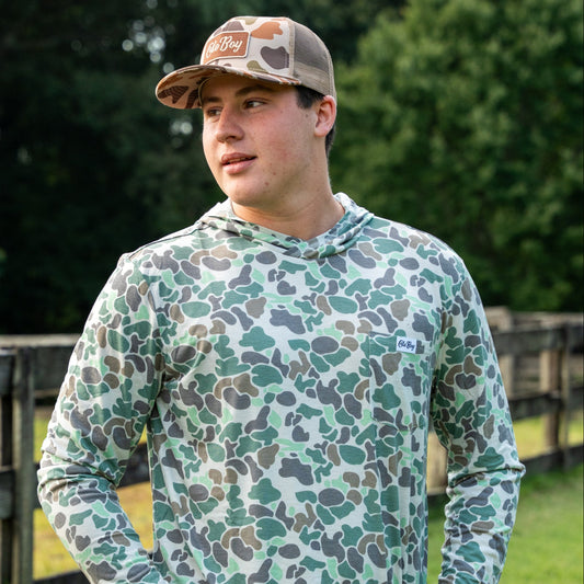 Man wearing a camouflage hoodie and cap standing outdoors with greenery in the background