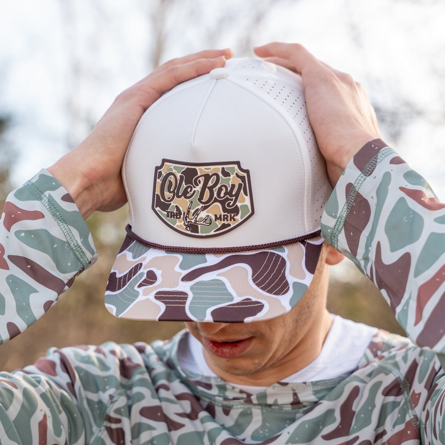 Person wearing a white cap with a logo and camouflage-patterned shirt outdoors