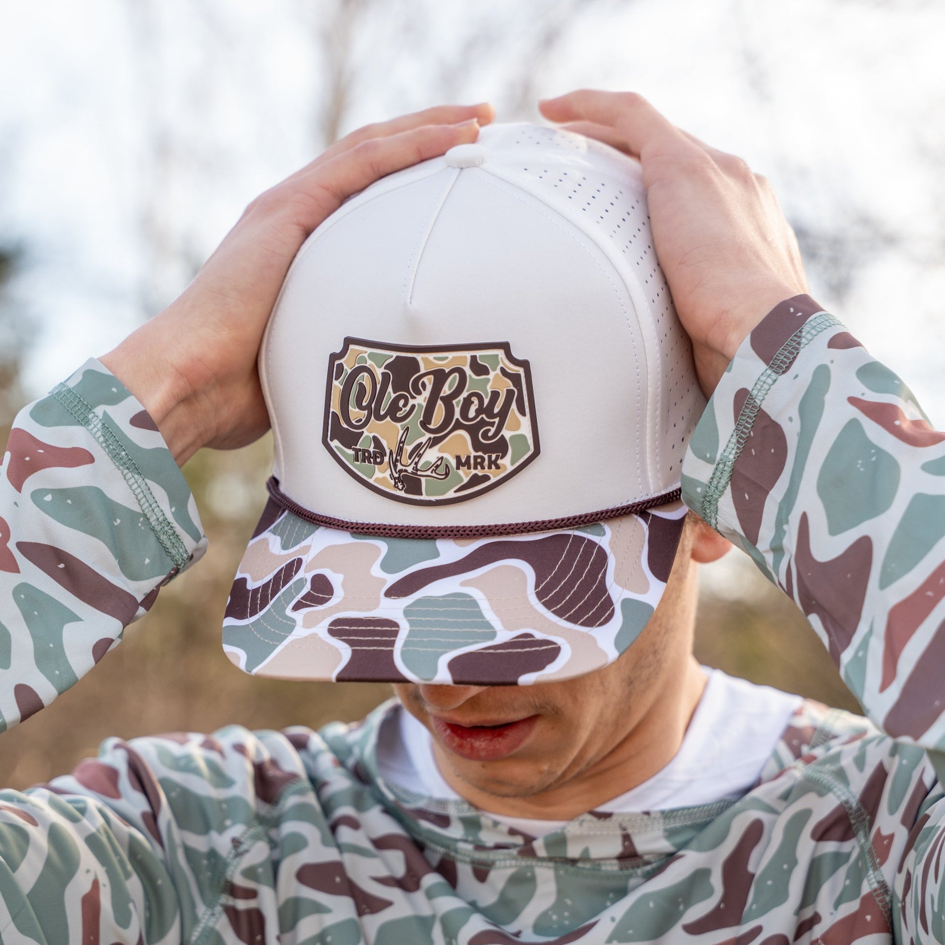 Person wearing a white cap with a logo and camouflage-patterned shirt outdoors